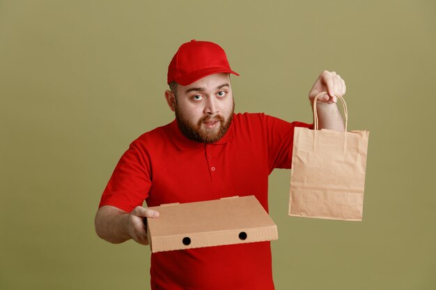 Delivery man employee in red cap blank tshirt uniform holding pizza box and paper bag looking at camera with serious face standing over green background