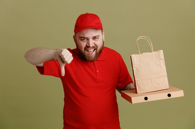 ピザの箱と紙袋を保持している赤い帽子の空白のTシャツの制服を着た配達人の従業員は、緑の背景の上に立っている親指を示すイライラした表情で叫んでカメラを見て