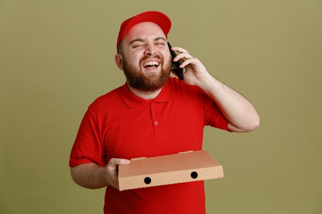 Delivery man employee in red cap blank tshirt uniform holding pizza box talking on mobile phone happy and excited laughing out standing over green background