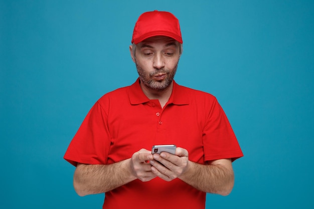 Free photo delivery man employee in red cap blank tshirt uniform holding smartphone looking at it being skeptic standing over blue background