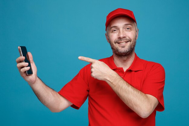 Сотрудник службы доставки в красной кепке с пустой футболкой держит смартфон, указывая на него указательным пальцем, смотрит в камеру с улыбкой на счастливом лице, стоящем на синем фоне