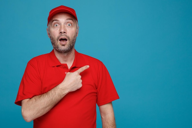 Delivery man employee in red cap blank tshirt uniform looking at camera amazed and surprised pointing with index finger to the side standing over blue background