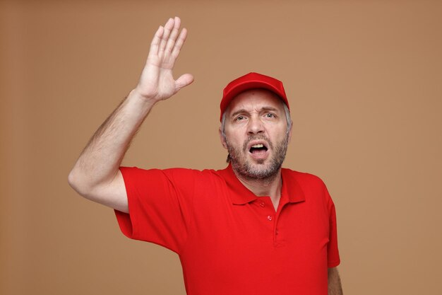 Delivery man employee in red cap blank tshirt uniform looking at camera angry and frustrated yelling raising arm standing over brown background