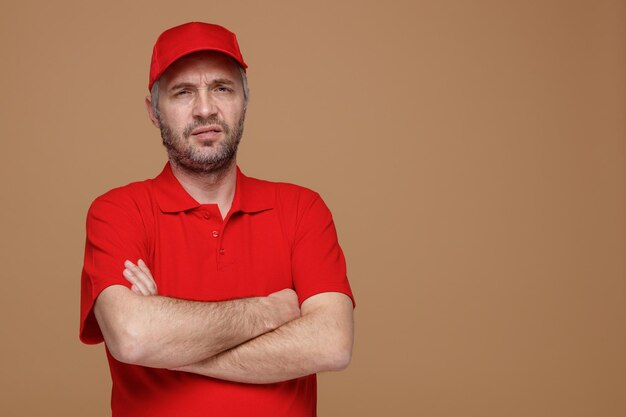 Delivery man employee in red cap blank tshirt uniform looking at camera being displeased offended with arms crossed on his chest standing over brown background