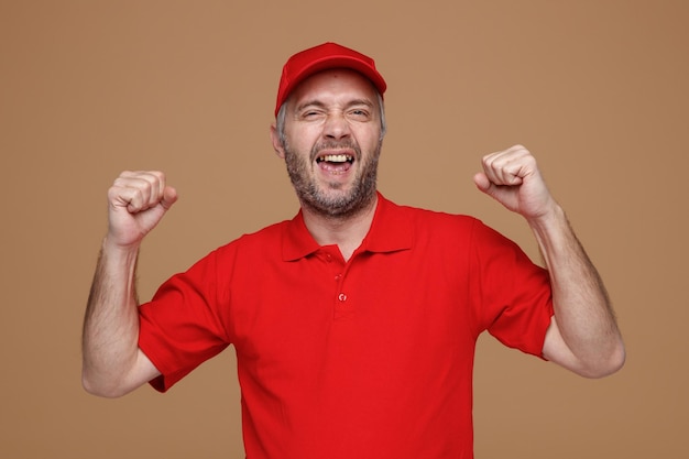 Сотрудник доставщика в красной кепке с пустой футболкой смотрит в камеру, безумно счастливый и взволнованный, поднимая сжатые кулаки, радуясь своему успеху, стоя на коричневом фоне