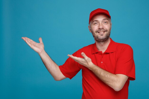 赤い帽子の空白のTシャツの制服を着た配達人の従業員がカメラを見て幸せで前向きな笑顔を元気に彼の手の腕で青い背景の上に立っている何かを提示