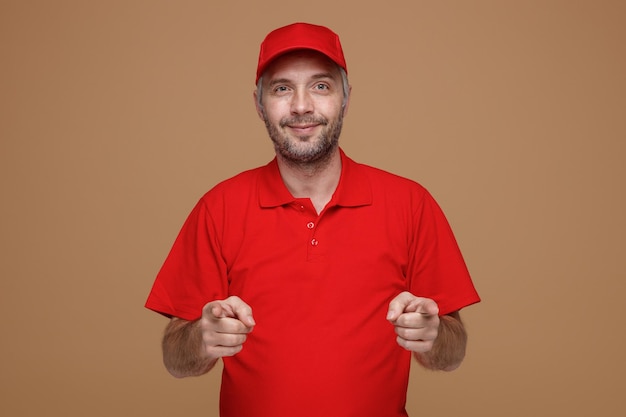 Сотрудник доставщика в красной кепке, пустой футболке, смотрит в камеру, счастливо и позитивно улыбаясь, указывая указательными пальцами обеими руками на камеру, стоящую на коричневом фоне