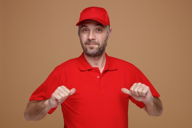 빨간 모자 빈 티셔츠 유니폼을 입은 배달원 직원이 갈색 배경 위에 서 있는 자신을 가리키며 만족스러운 미소를 지으며 카메라를 바라보고 있습니다.