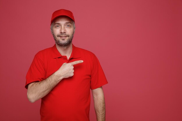 赤い帽子の空白のTシャツの制服を着た配達人の従業員は、赤い背景の上に立っている側に人差し指でフレンドリーなポインティングを笑顔でカメラを見て