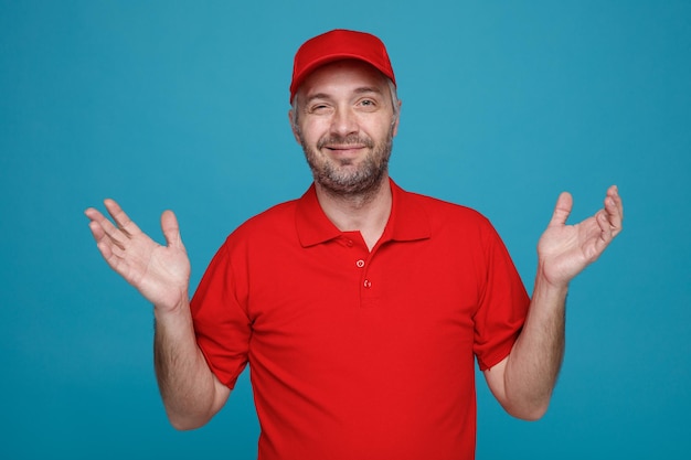 Dipendente uomo di consegna in uniforme tshirt bianca con cappuccio rosso guardando la fotocamera sorridente amichevole alzando le braccia in piedi su sfondo blu