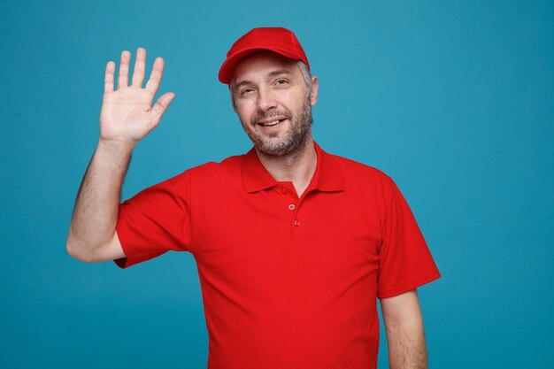 Delivery man employee in red cap blank tshirt uniform looking at camera smiling friendly waving with hand standing over blue background