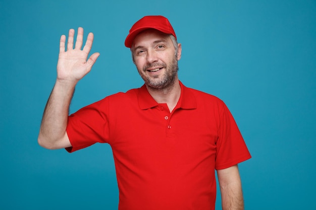 빨간 모자 빈 티셔츠를 입은 배달원 직원이 파란 배경 위에 손을 들고 친절하게 손을 흔들며 웃고 있는 카메라를 바라보고 있습니다.
