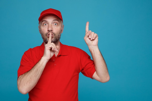 カメラを見て驚いた赤い帽子の空白のTシャツの制服を着た配達人の従業員は、青い背景の上に立っている人差し指で指している唇に指で沈黙のジェスチャーをしている