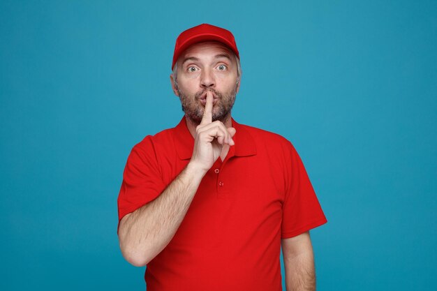赤い帽子の空白のTシャツの制服を着た配達人の従業員がカメラを見て驚いた青い背景の上に立っている唇に指で沈黙のジェスチャーをする