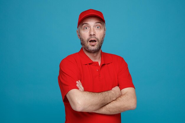 Delivery man employee in red cap blank tshirt uniform looking at camera surprised with crossed arms on his chest standing over blue background