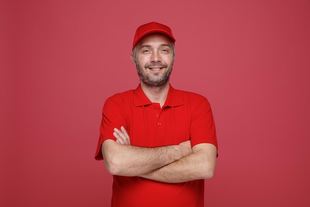 Delivery man employee in red cap blank tshirt uniform looking at camera with arms crossed on his chest smiling cheerfully expression standing over red background