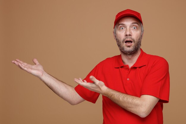 カメラを見て赤いキャップの空白のTシャツの制服を着た配達人の従業員は、茶色の背景の上に立っている何かを彼の手の腕で提示して驚いて驚いた