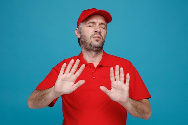Delivery man employee in red cap blank tshirt uniform looking displeased making defense gesture refusing standing over blue background