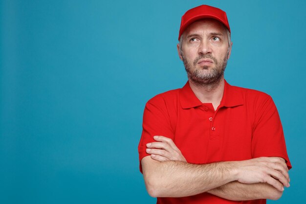 青い背景の上に立っている彼の胸に腕を組んで不機嫌になって見上げている赤い帽子の空白のTシャツの制服を着た配達人の従業員