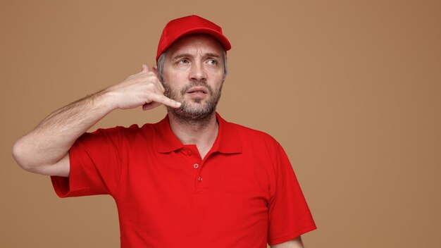 Delivery man employee in red cap blank tshirt uniform making call me gesture looking aside being offended and displeased standing over brown background