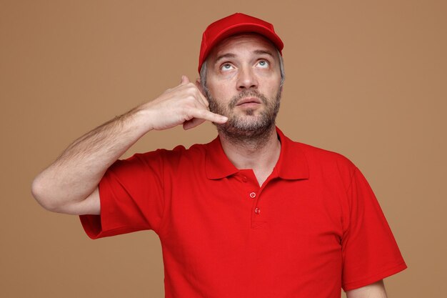 Delivery man employee in red cap blank tshirt uniform making call me gesture looking up confused thinking standing over brown background