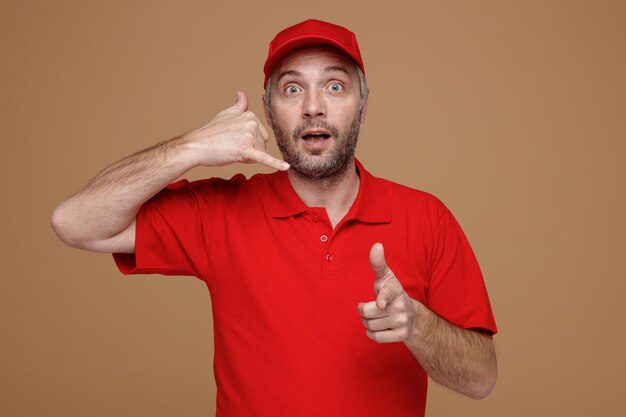 Сотрудник доставщика в красной кепке с пустой футболкой делает жест, звонит мне, указывая указательным пальцем на камеру, счастливый и удивленный, стоя на коричневом фоне