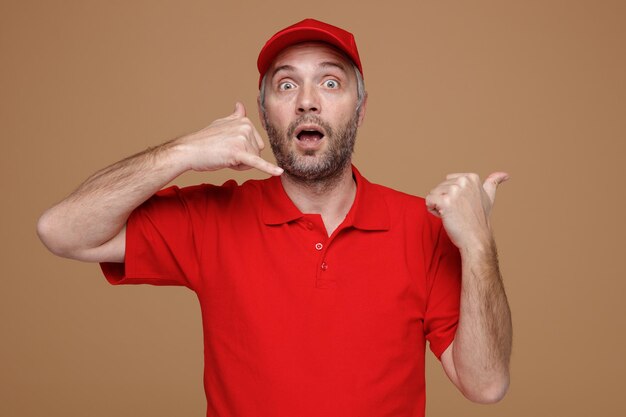 Delivery man employee in red cap blank tshirt uniform making call me gesture pointing with thumb to the side looking surprised and amazed standing over brown background