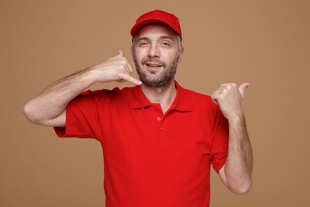 Delivery man employee in red cap blank tshirt uniform making call me gesture pointing with thumb to the side smiling friendly happy and positive standing over brown background