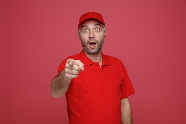 빨간 모자 빈 티셔츠를 입은 배달원 직원이 집게 손가락으로 카메라를 가리키며 빨간색 배경 위에 서 있는 놀란 표정을 하고 있다