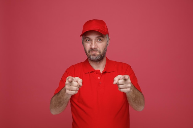 Dipendente uomo di consegna in uniforme tshirt bianca con cappuccio rosso che punta con il dito indice alla fotocamera sorridente amichevole felice e positivo in piedi su sfondo rosso