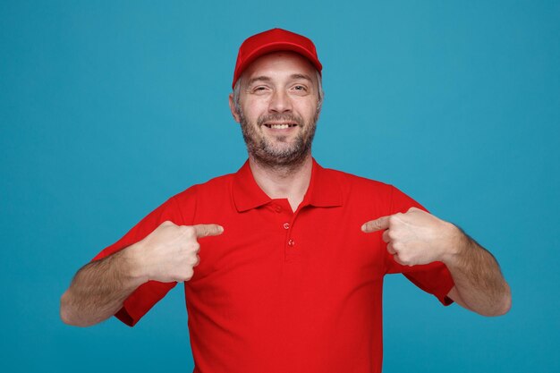 赤い帽子の空白のTシャツの制服を着た配達人の従業員は、人差し指で自分自身を指して、青い背景の上に立って幸せで幸せなカメラを見て自信を持って笑っています