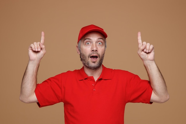 Delivery man employee in red cap blank tshirt uniform pointing with index fingers up looking at camera surprised standing over brown background