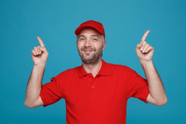 검지 손가락으로 가리키는 빨간색 모자 빈 티셔츠 유니폼을 입은 배달원 직원은 파란색 배경 위에 서 있는 카메라를 보며 자신감 있게 웃고 있습니다.