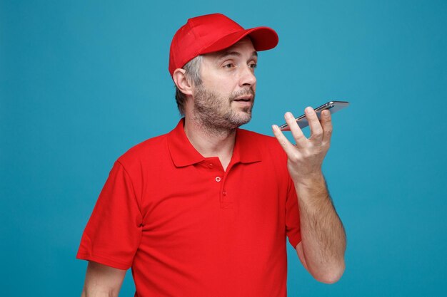 Delivery man employee in red cap blank tshirt uniform recording voice message using smartphone looking confident standing over blue background