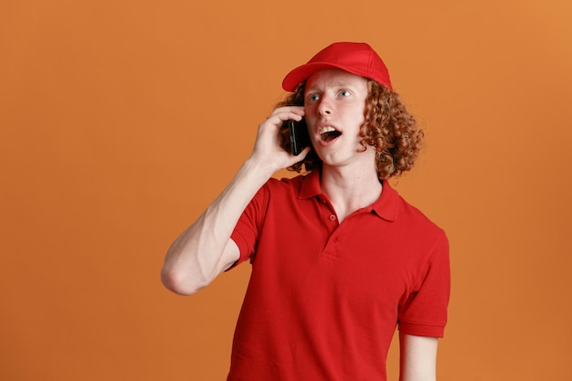 Dipendente dell'uomo delle consegne in uniforme della maglietta vuota con cappuccio rosso che parla al telefono cellulare che sembra stupito e sorpreso in piedi su sfondo arancione