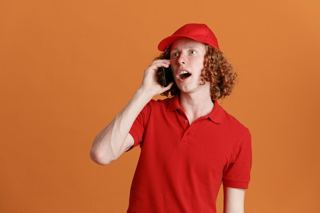 Сотрудник службы доставки в красной кепке с пустой футболкой разговаривает по мобильному телефону, выглядя изумленным и удивленным, стоя на оранжевом фоне