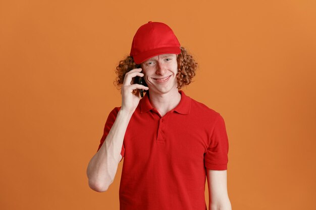 赤い帽子の空白のTシャツの制服を着た配達人の従業員は、オレンジ色の背景の上にフレンドリーで幸せで前向きに立って笑顔のカメラを見て携帯電話で話しています