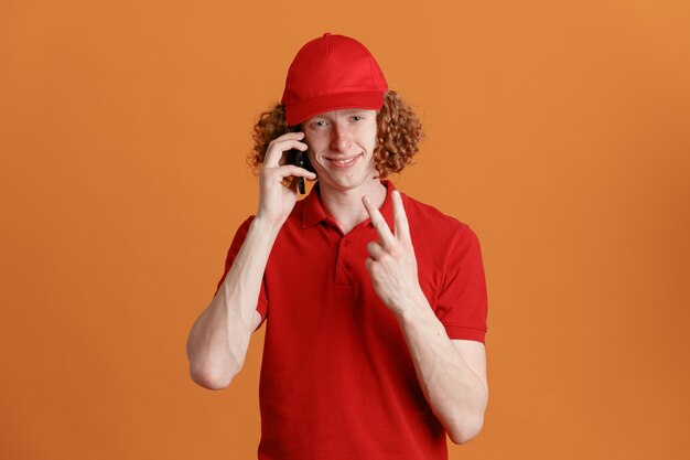 빨간 모자를 쓴 빈 티셔츠를 입은 배달원 직원이 주황색 배경 위에 서 있는 2번 또는 vsign을 보여주는 카메라를 보며 휴대폰 통화를 하고 있다