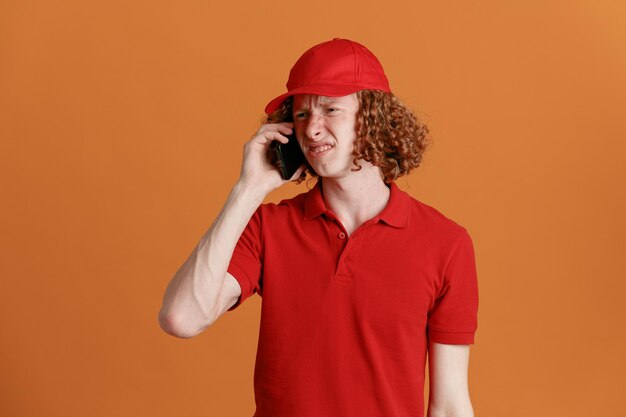 Сотрудник доставщика в красной кепке с пустой футболкой разговаривает по мобильному телефону, выглядя недовольным, кривя рот, стоя на оранжевом фоне