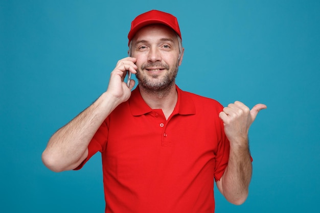 Сотрудник доставщика в красной кепке, пустой футболке, разговаривает по мобильному телефону, улыбается счастливо и позитивно, указывая большим пальцем в сторону, стоя на синем фоне