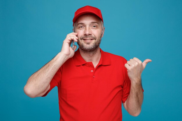 Сотрудник доставщика в красной кепке, пустой футболке, разговаривает по мобильному телефону, улыбается счастливо и позитивно, указывая большим пальцем в сторону, стоя на синем фоне