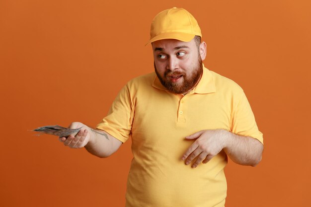 Delivery man employee in yellow cap blank tshirt uniform holding cash offering to someone looing happy and positive standing over orange background