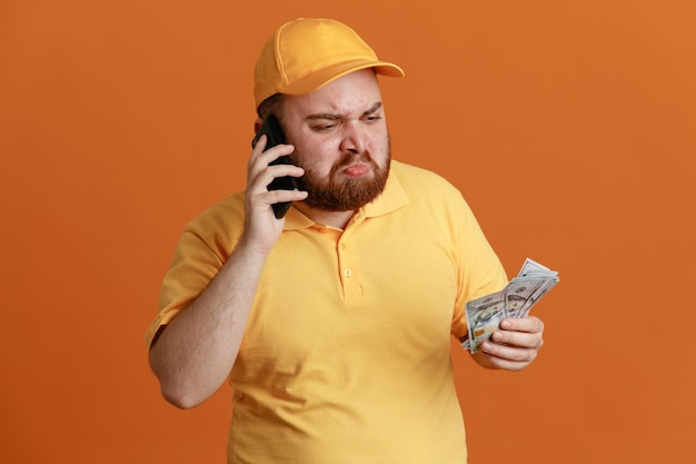 Delivery man employee in yellow cap blank tshirt uniform holding cash talking on mobile phone being upset with sad expression standing over orange background