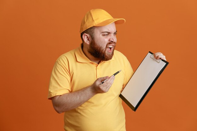 Сотрудник службы доставки в желтой кепке с пустой футболкой держит планшет и ручку, глядя в сторону, злой и разочарованный крича, стоя на оранжевом фоне