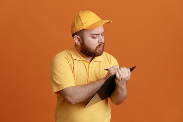 Free photo delivery man employee in yellow cap blank tshirt uniform holding clipboard and pen making notes looking confident standing over orange background