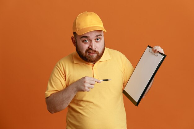 Delivery man employee in yellow cap blank tshirt uniform holding clipboard and pen pointing with it at clipboard looking at camera smiling standing over orange background