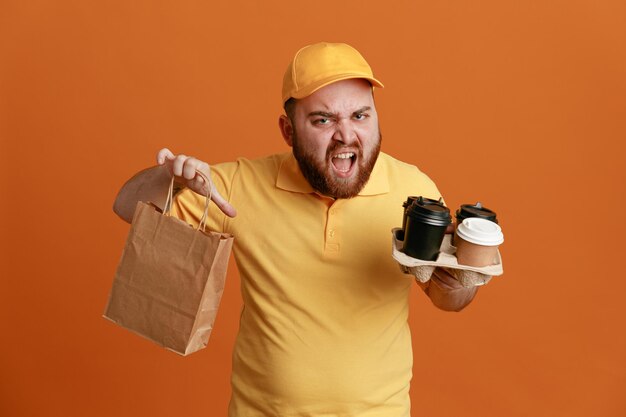 コーヒーカップと紙袋を保持している黄色いキャップの空白のTシャツの制服を着た配達人の従業員は、オレンジ色の背景の上に立って怒って欲求不満の叫び声を上げています