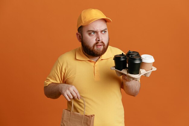 Delivery man employee in yellow cap blank tshirt uniform holding coffee cups and paper bag looking aside with frowning face standing over orange background