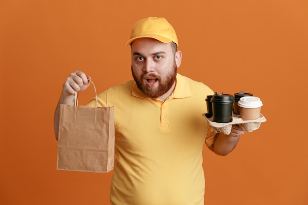 オレンジ色のキャップの空白のTシャツの制服を着た配達人の従業員は、オレンジ色の背景の上に立って幸せで驚きのカメラを見てコーヒーカップと紙袋を保持しています