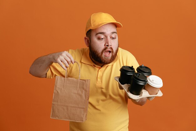 Сотрудник доставщика в желтой кепке с пустой футболкой, держащий кофейные чашки и бумажный пакет, выглядит смущенным и удивленным, стоя на оранжевом фоне